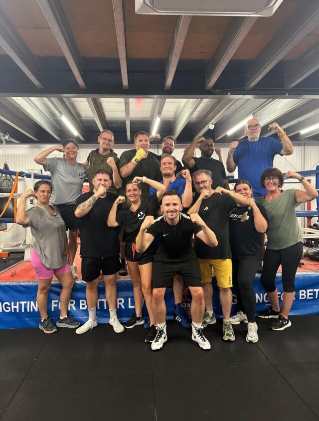 Fifteen participants standing in front of a boxing ring at Boxing Futures in Yaxley, all flexing their arms and smiling after a CDA deaf boxing session.