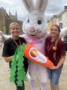 Peterborough United mascot Peter Burrow holding a carrot branded with the Cambridgeshire Deaf Association logo, alongside CDA staff members Sanita Salineice and Becky Fitzgerald