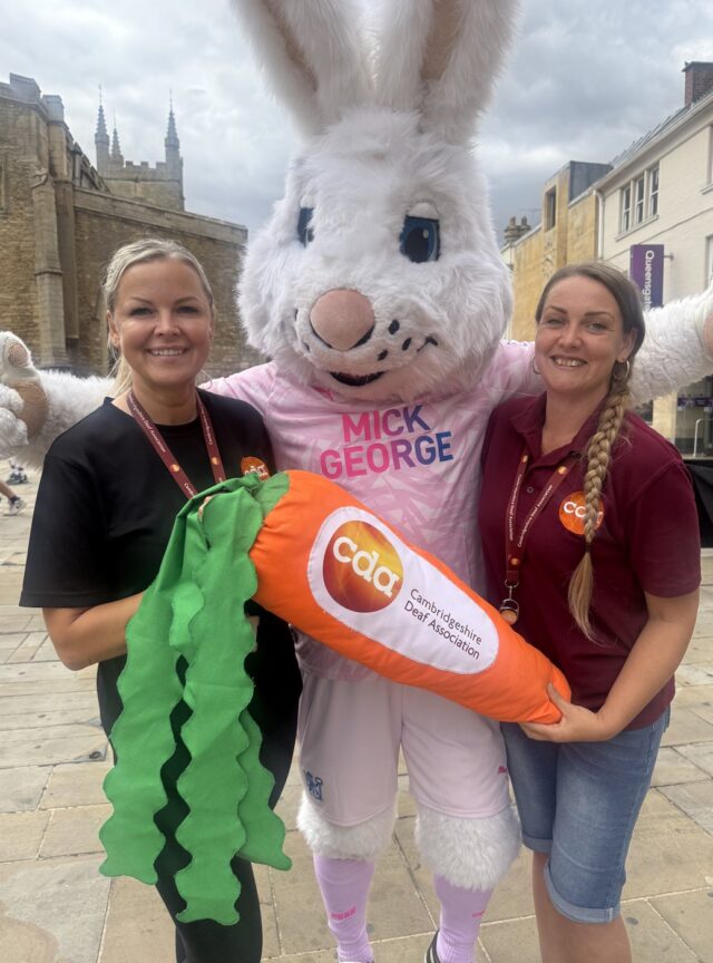 Peterborough United mascot Peter Burrow holding a carrot branded with the Cambridgeshire Deaf Association logo, alongside CDA staff members Sanita Salineice and Becky Fitzgerald