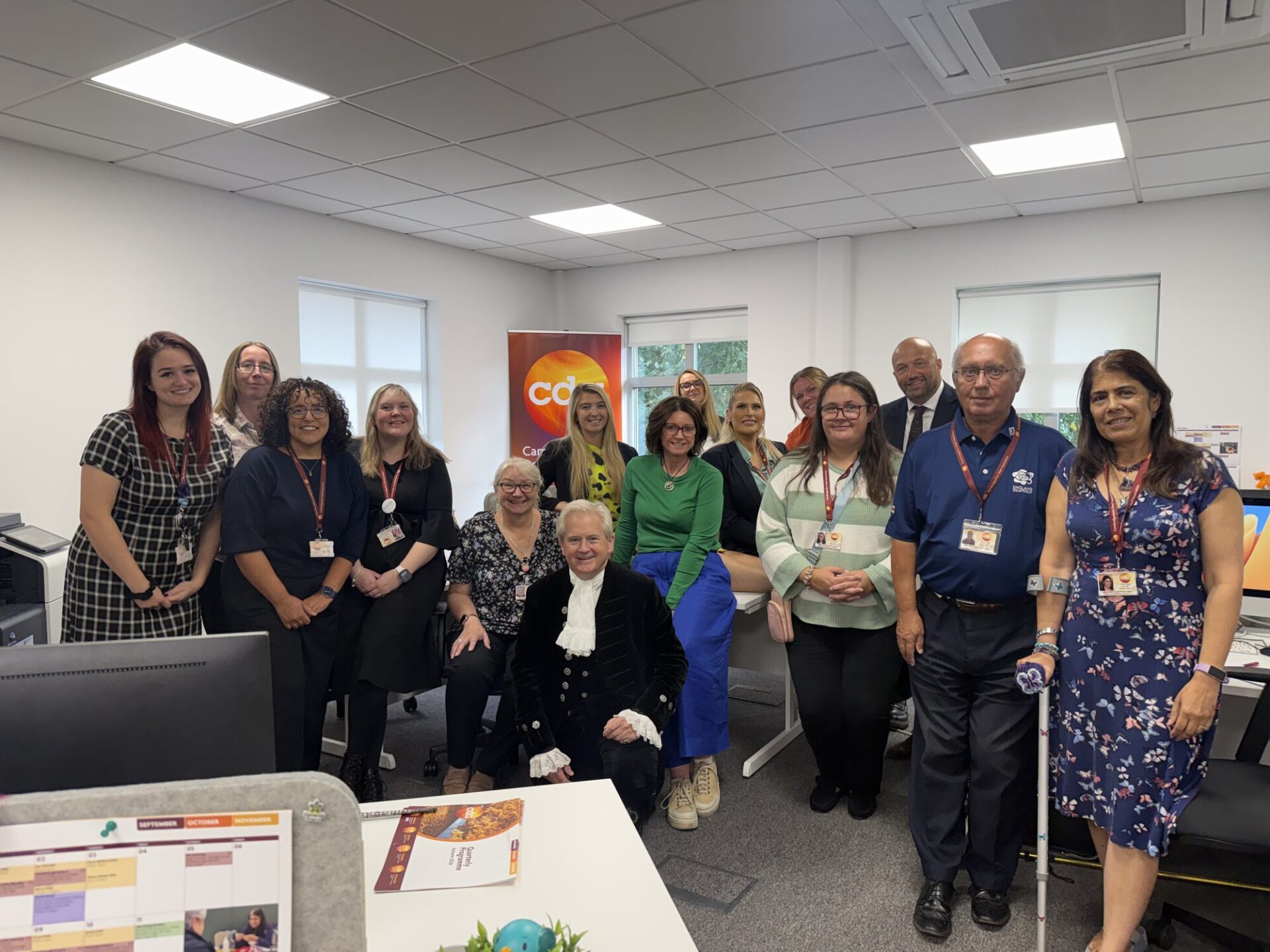 CDA staff and volunteers gathered in the Huntingdon office with the High Sheriff of Cambridgeshire, who is kneeling at the front in court dress, with a CDA banner behind them