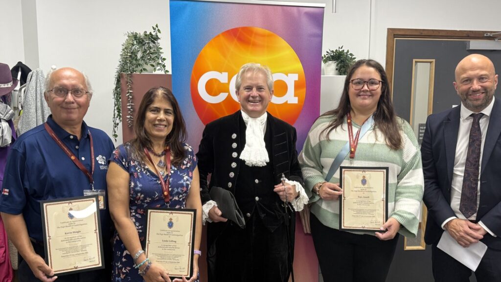 Volunteers Kevin Hingle, Linda Gilling and Faye Axcell holding their framed High Sheriff's Certificates of Appreciation, standing with the High Sheriff of Cambridgeshire in court dress and CDA Chief Executive Andrew Palmer in front of a CDA banner