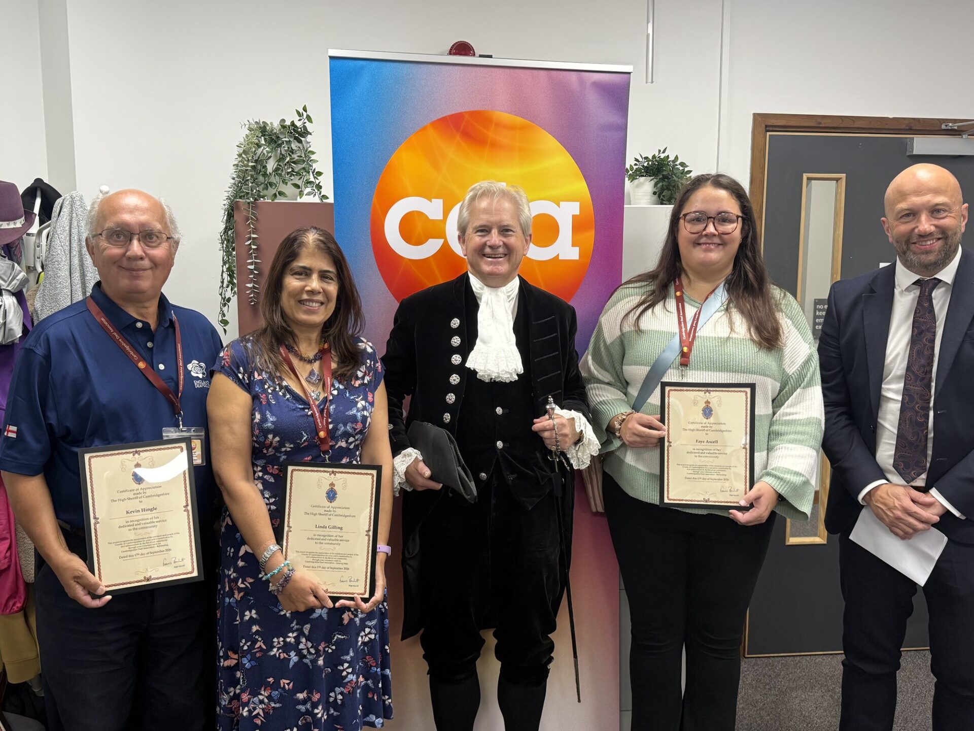 Volunteers Kevin Hingle and Linda Gilling, the High Sheriff of Cambridgeshire in court dress, volunteer Faye Axcell and CDA Chief Executive Andrew Palmer. The three volunteers hold framed Certificates of Appreciation in front of a CDA banner