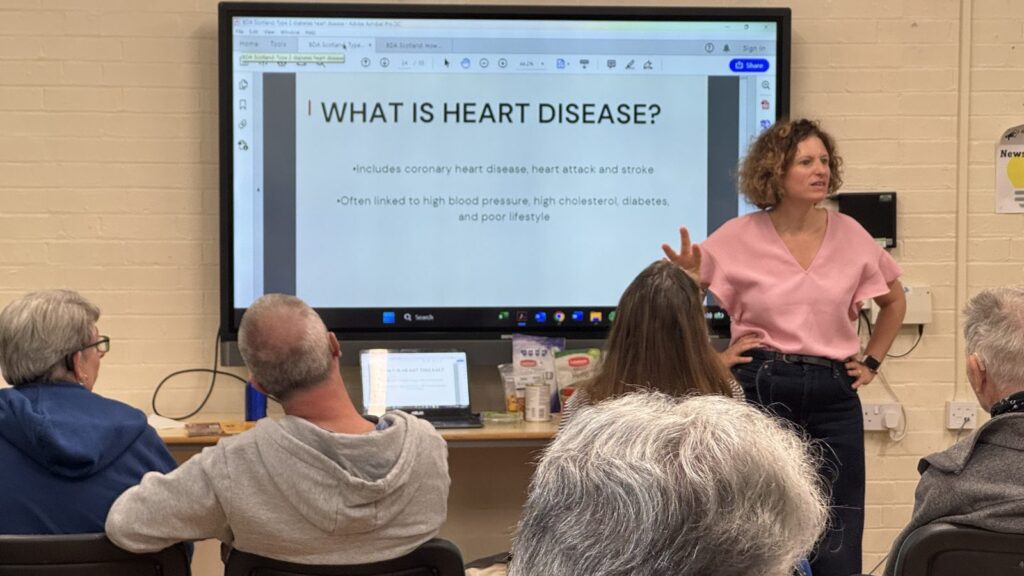 Nutritional therapist Naomi Goldman signing to an audience at the Peterborough Deaf Drop-In, in front of a slide reading What is heart disease?